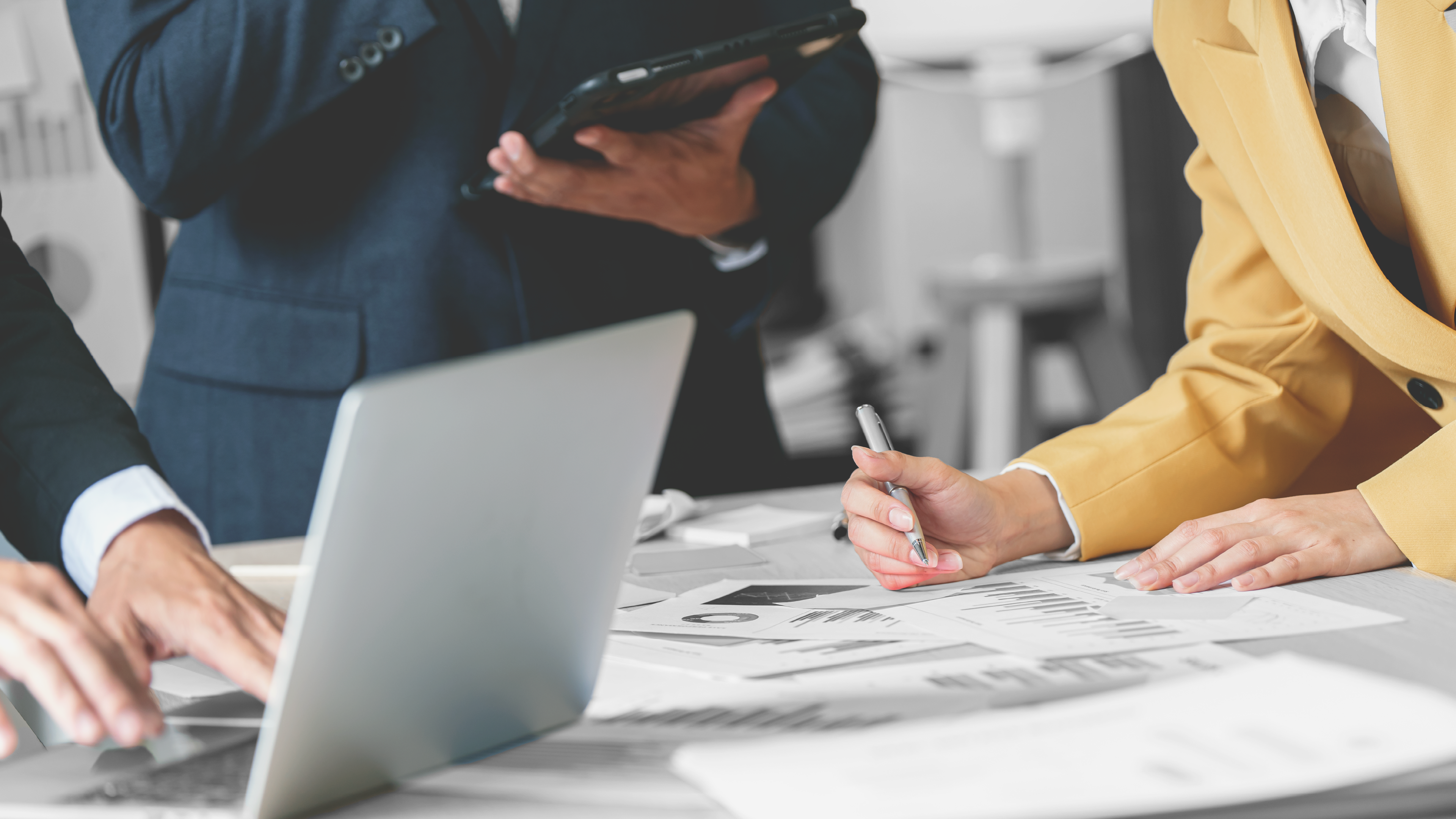 Imagen de tres personas trabajando: una en computadora, otra con tableta y una mujer tomando notas a mano