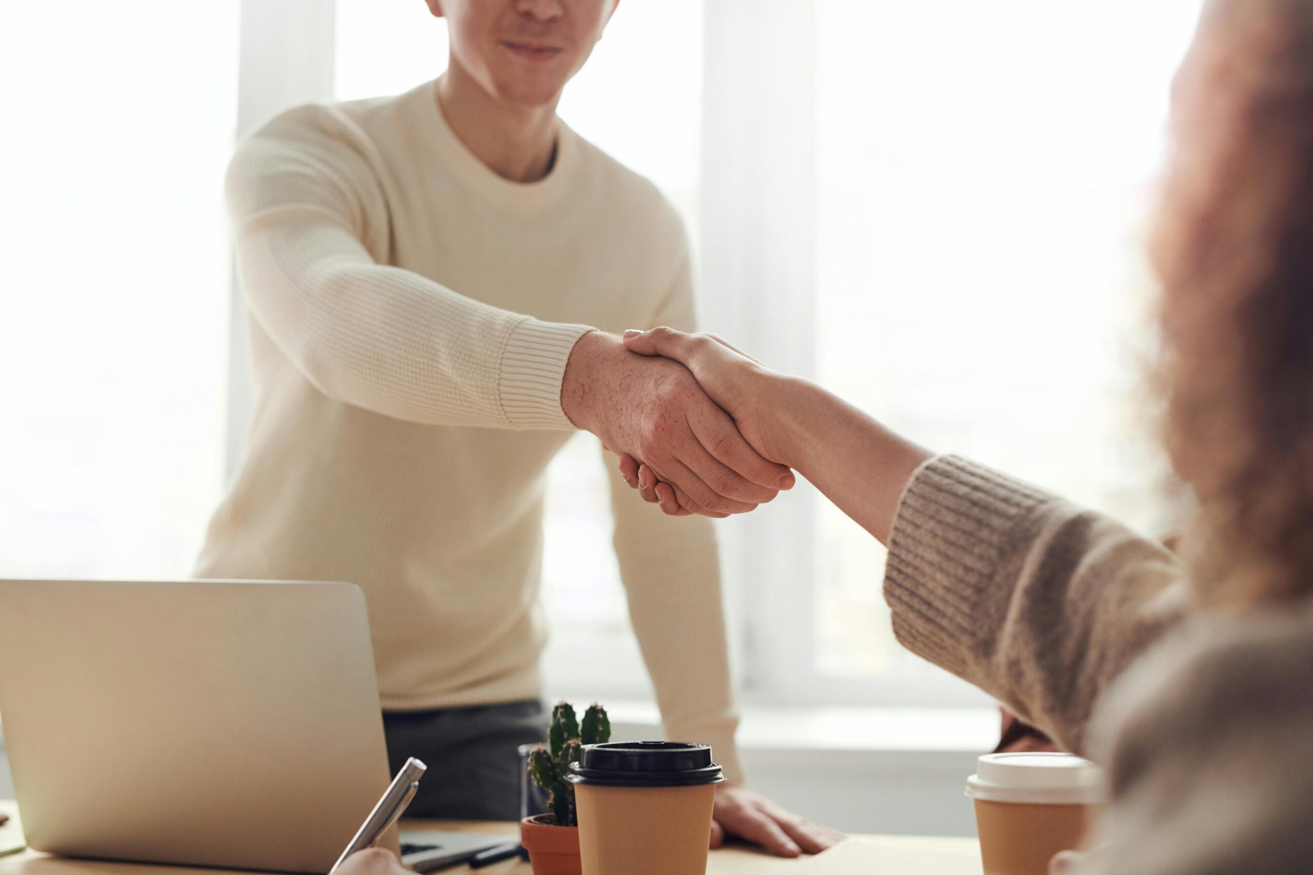 Imagen de dos personas dándose la mano en una oficina, con una computadora portátil y una taza de café en la mesa